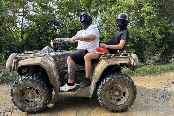 Two people ride a muddy ATV on a dirt path, wearing helmets and sunglasses.