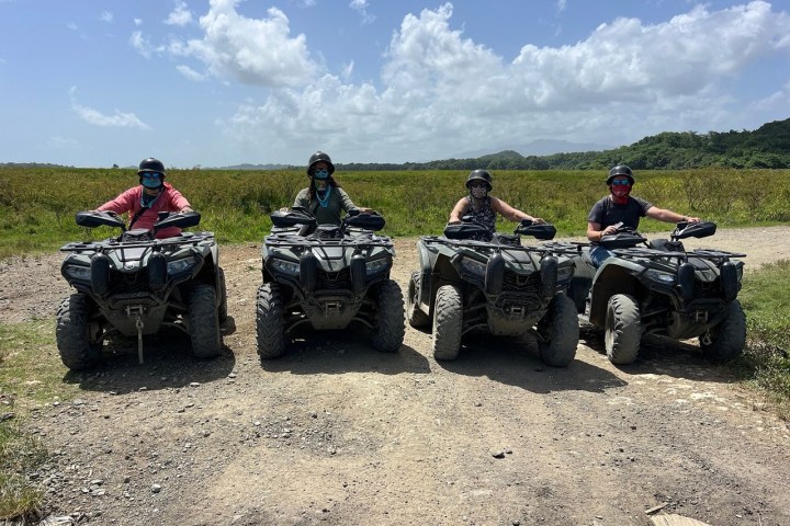 Four people in helmets on ATVs lined up on a dirt path with grass and hills in the background.