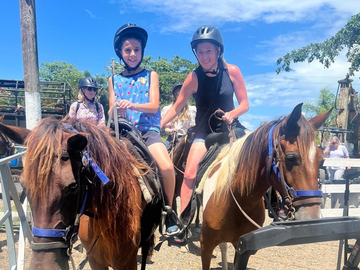 Two people wearing helmets riding horses outdoors on a sunny day.