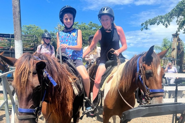 Two people wearing helmets riding horses outdoors on a sunny day.