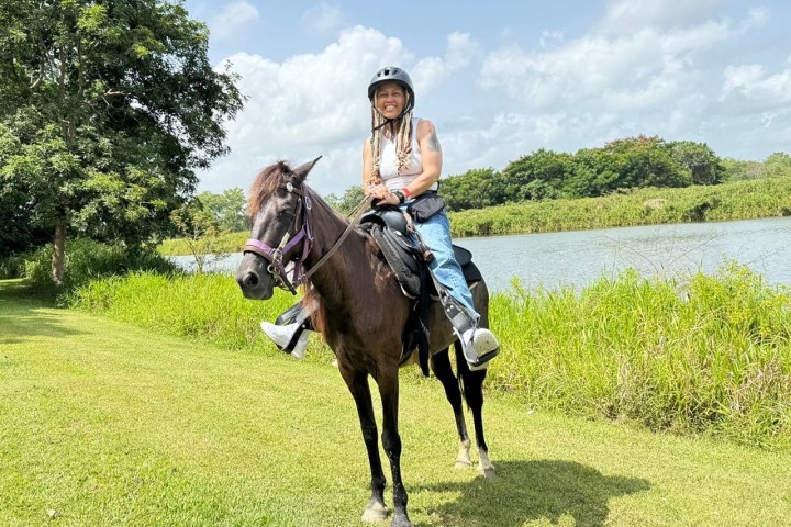 Person in helmet riding a horse near a lake, surrounded by grass and trees under a partly cloudy sky.