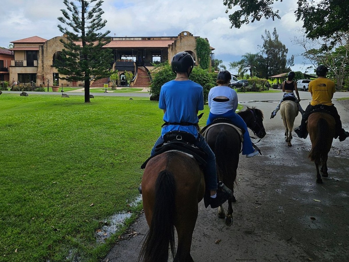 Group of people riding horses towards a large building on a grassy area.