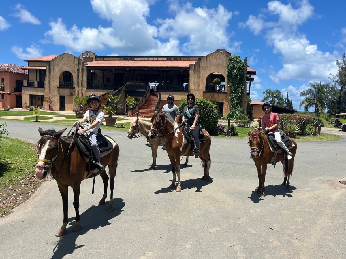 Four people on horseback outside a large, rustic building under a blue sky.