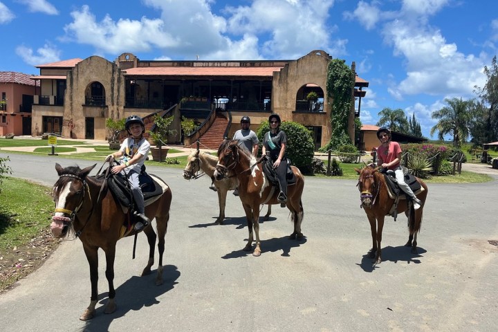 Four people on horseback outside a large, rustic building under a blue sky.