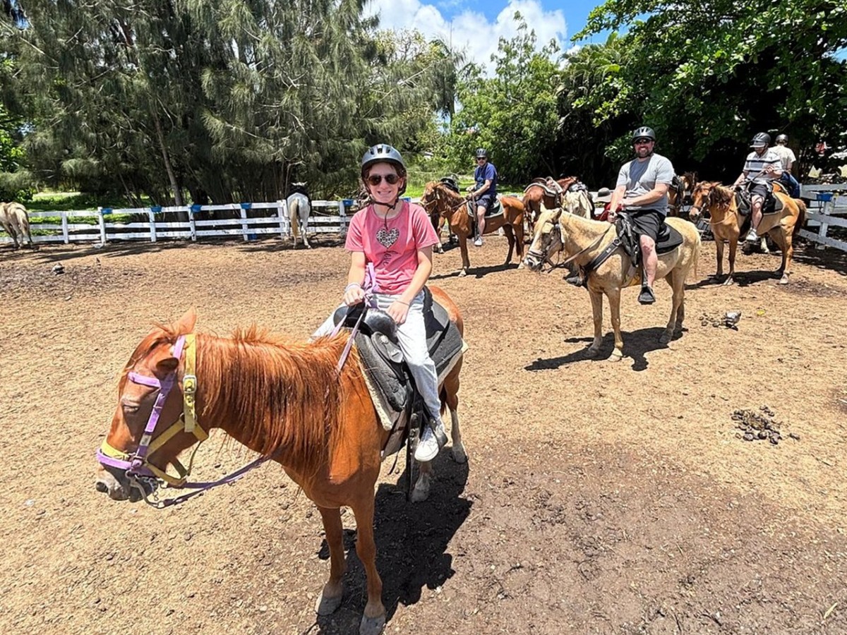 Group of people horseback riding in a sunny paddock, wearing helmets.