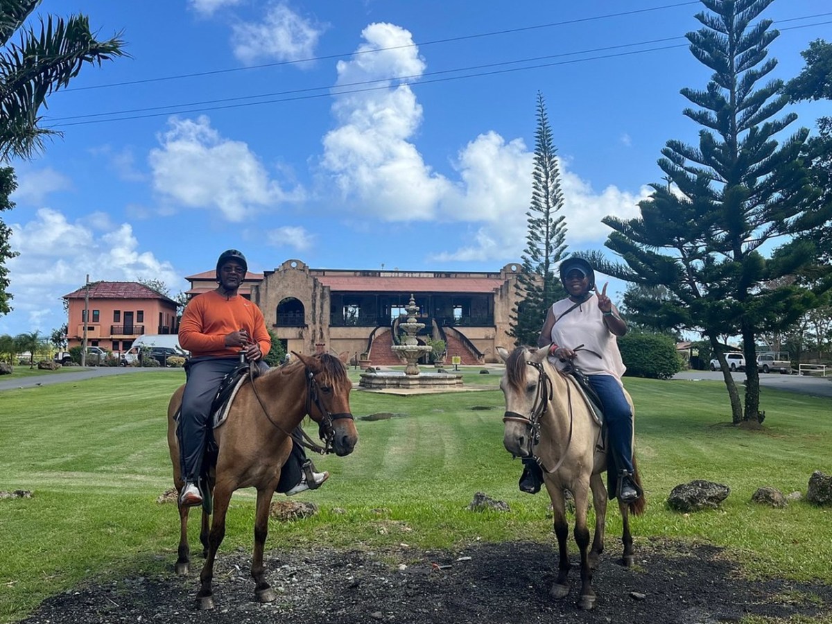 Two people on horseback in front of a large building and fountain with trees around.
