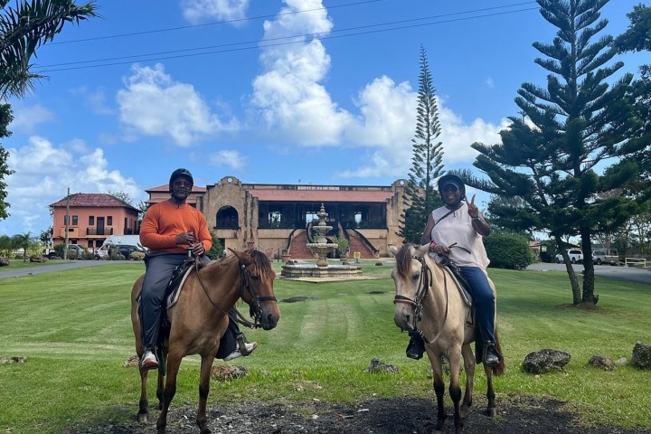 Two people on horseback in front of a large building and fountain with trees around.