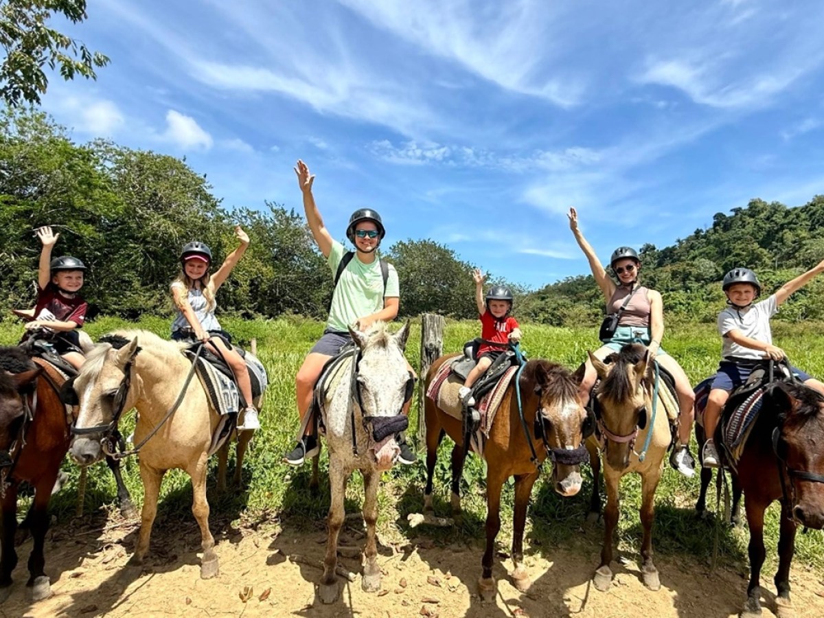 Six people riding horses, wearing helmets, waving in a lush countryside setting.