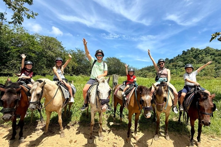 Six people riding horses, wearing helmets, waving in a lush countryside setting.