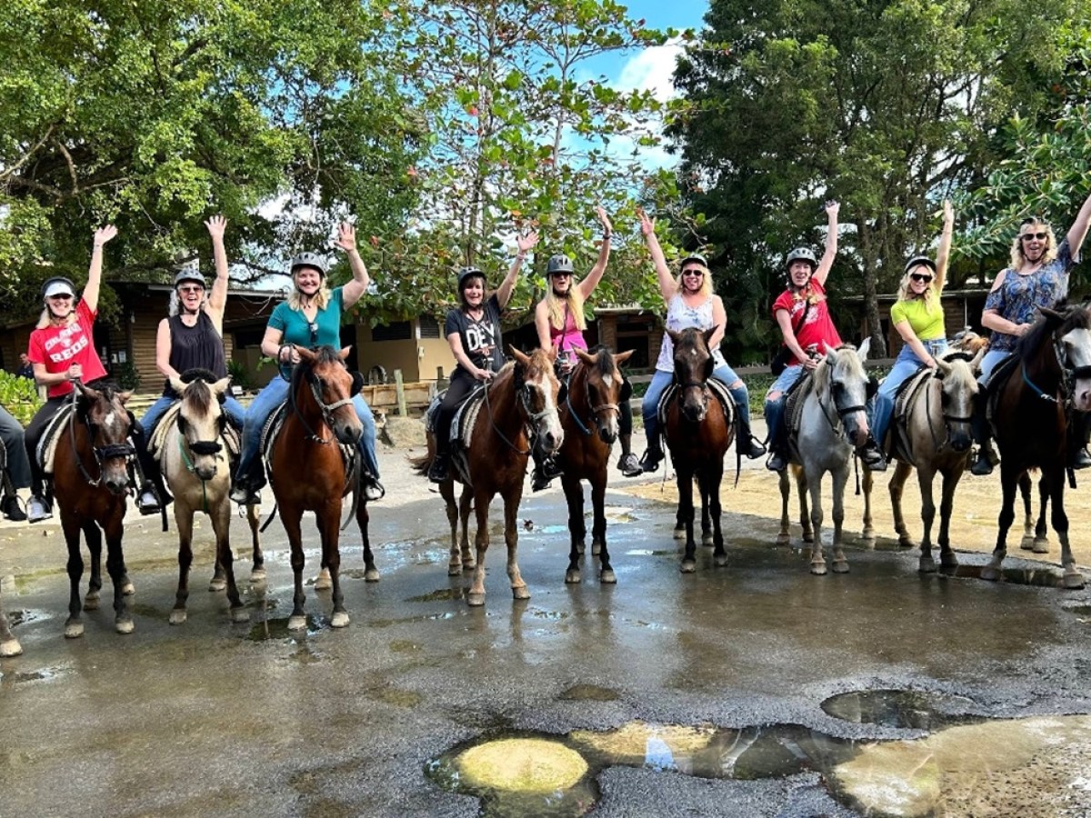A group of people on horseback raising arms, wearing helmets, in an outdoor setting with trees.