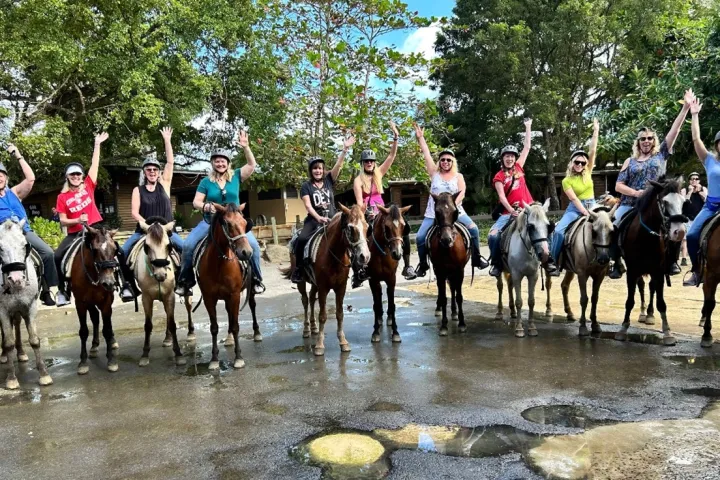 A group of people on horseback raising arms, wearing helmets, in an outdoor setting with trees.