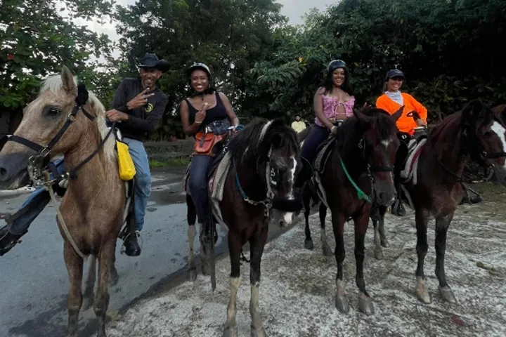 Four people riding horses on a path, surrounded by trees, with one person in a bright orange jacket.