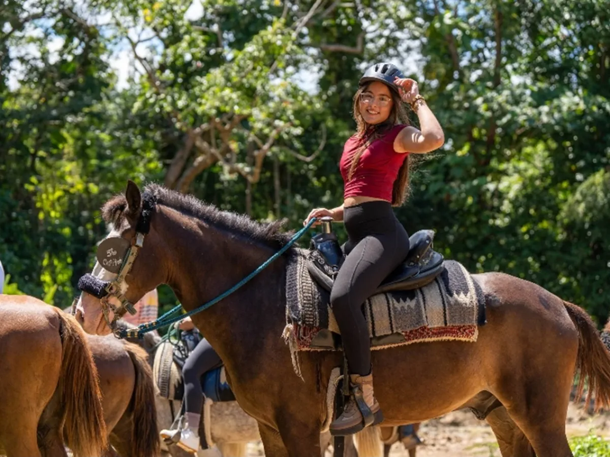 Person in helmet riding a horse, wearing a red top and black pants, with a forest background.