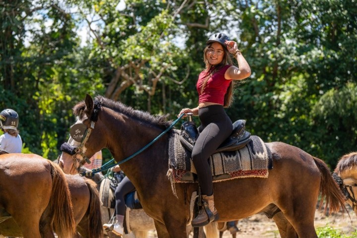 Person in helmet riding a horse, wearing a red top and black pants, with a forest background.