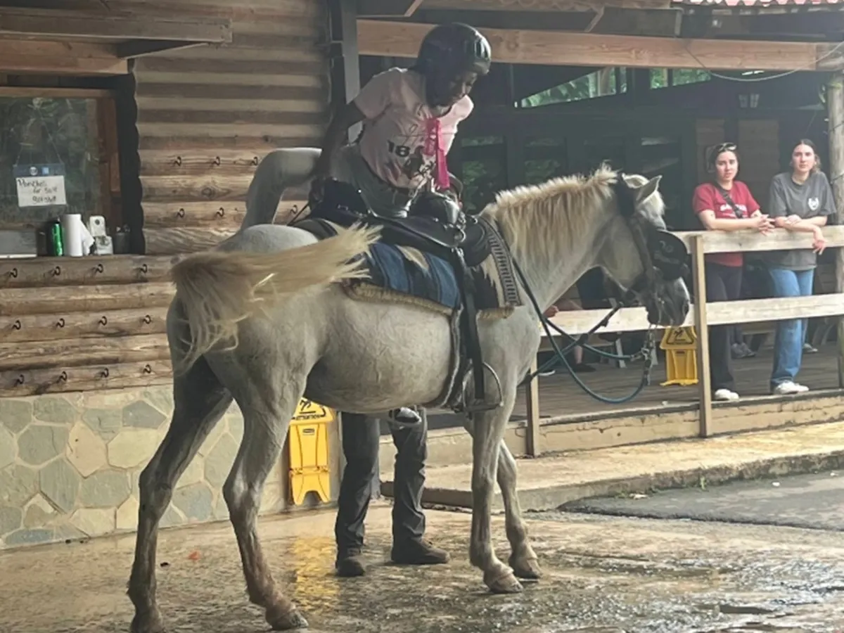 Person in helmet riding a white horse near a wooden building with onlookers in the background.