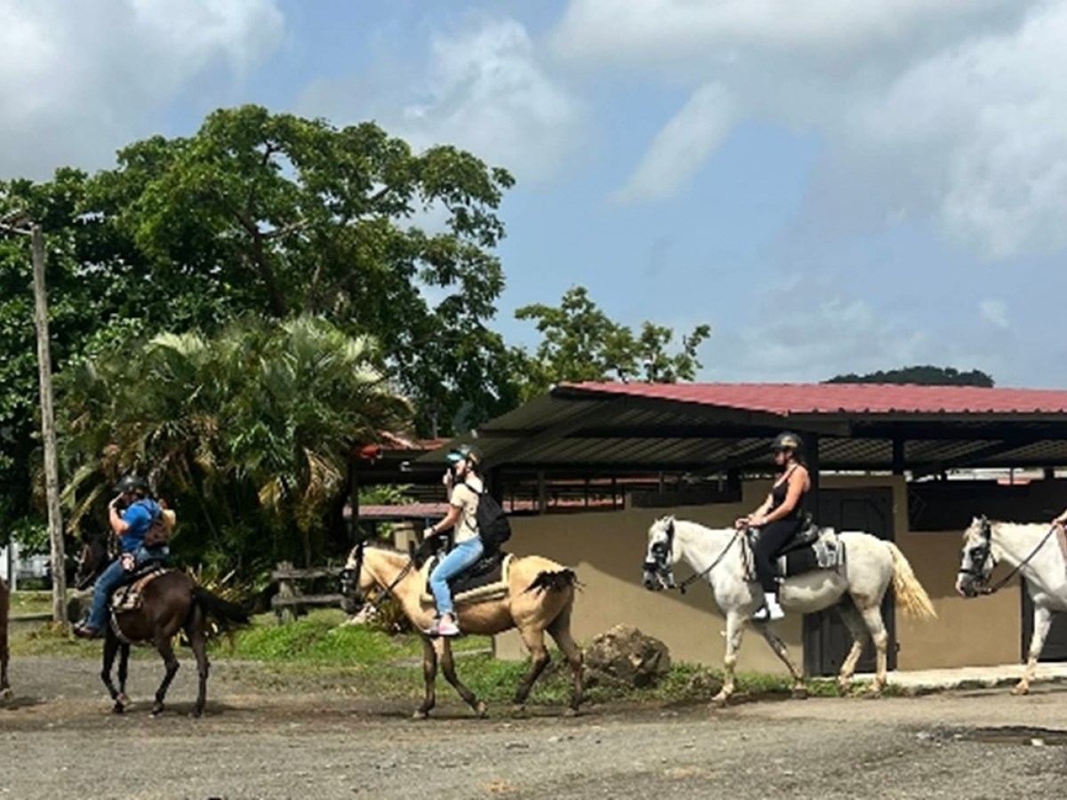 Group of people riding horses near a building on a sunny day.