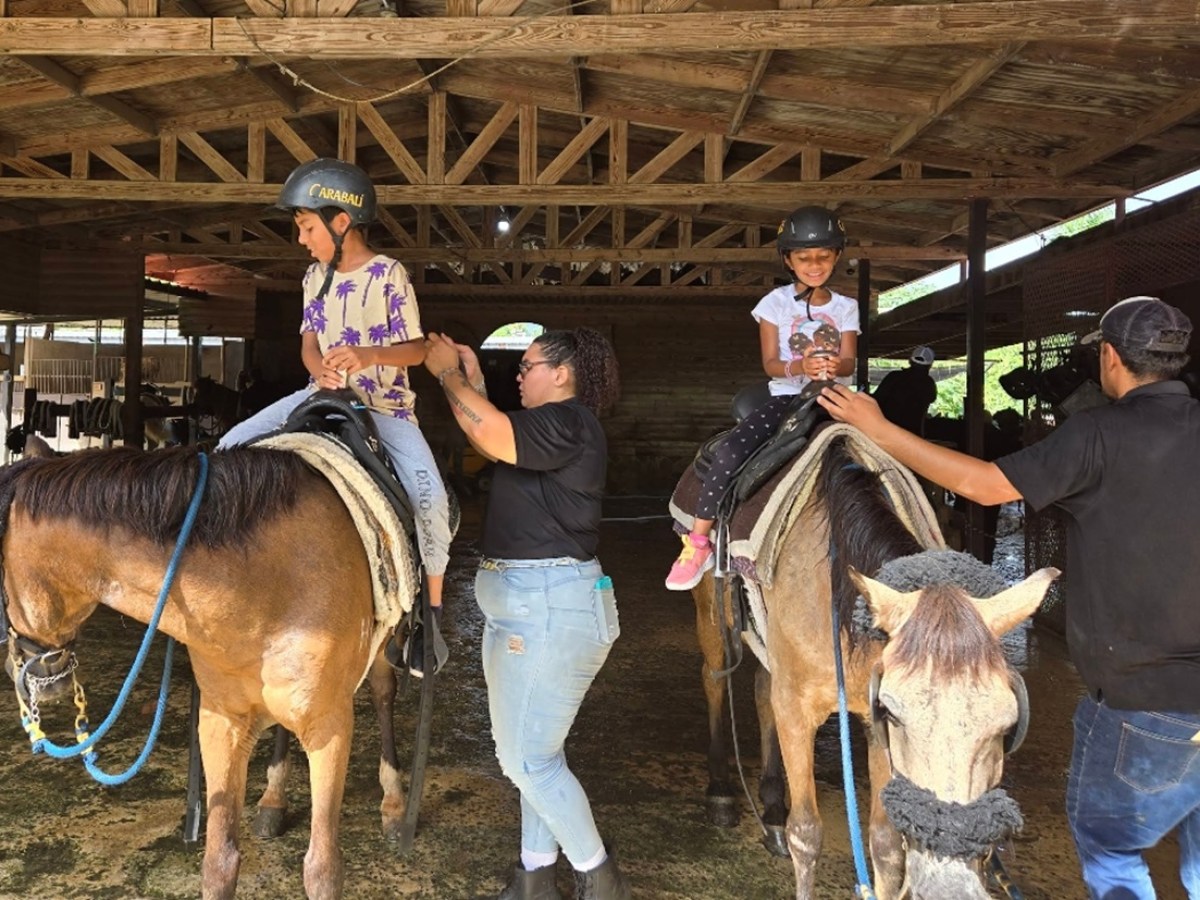 Two children on horses with helmets, assisted by adults in a wooden stable.