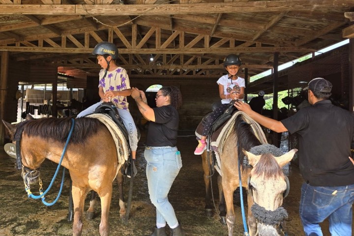 Two children on horses with helmets, assisted by adults in a wooden stable.