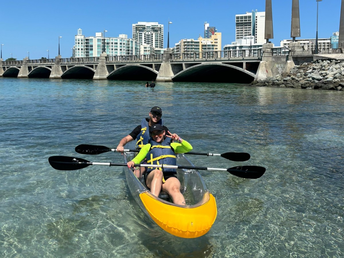 Two people in a clear kayak on calm water near a bridge and city skyline.