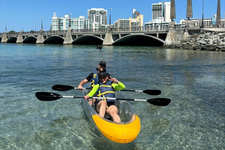 Two people in a clear kayak on calm water near a bridge and city skyline.