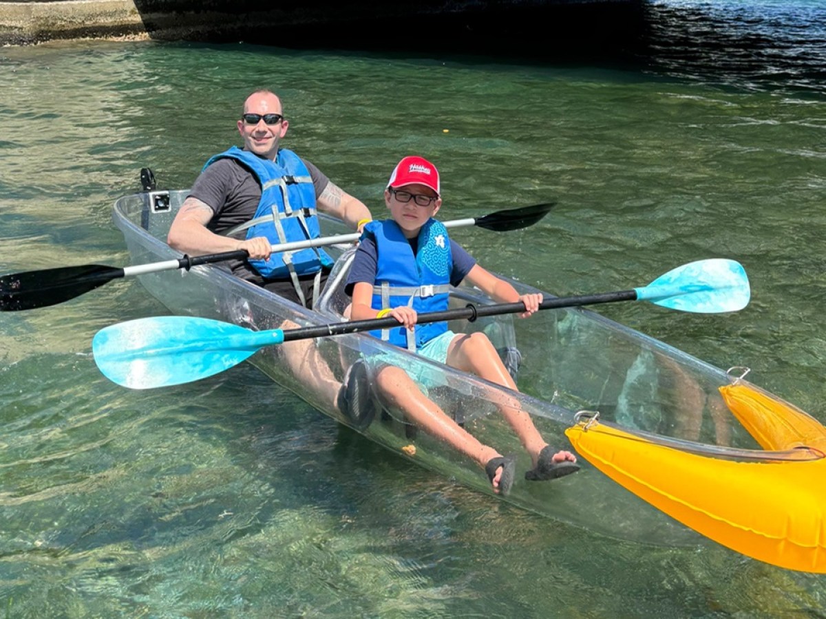 Two people kayaking in a clear kayak on calm water.