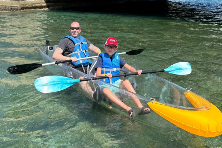 Two people kayaking in a clear kayak on calm water.