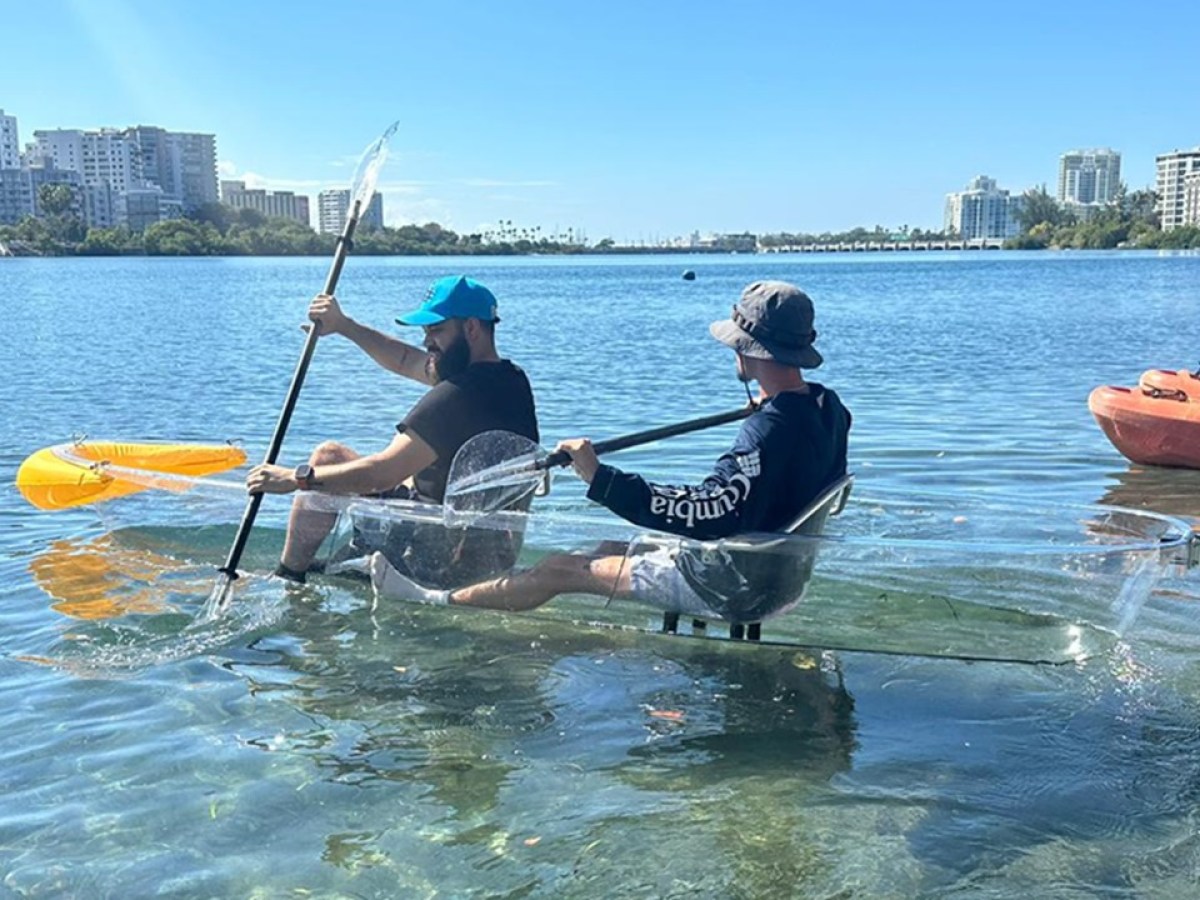 Two people kayaking in a clear kayak on a sunny day near a city waterfront.