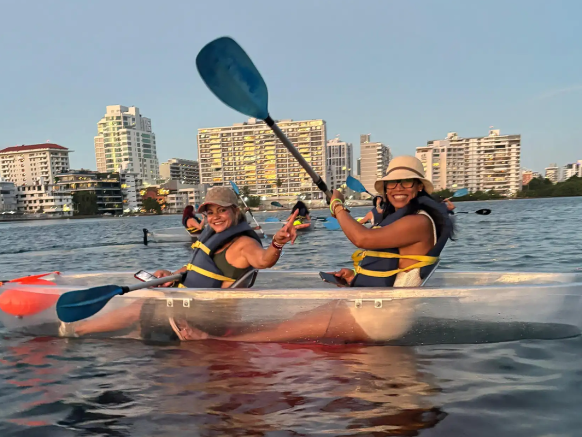 Two people in a transparent kayak on a city waterway, wearing life vests and hats.