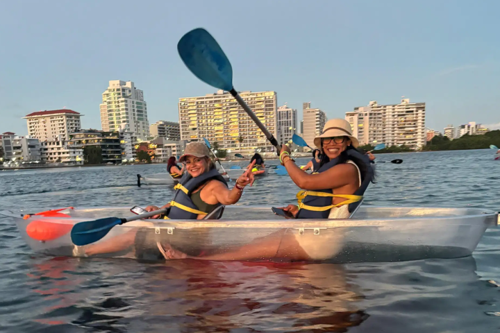 Two people in a transparent kayak on a city waterway, wearing life vests and hats.