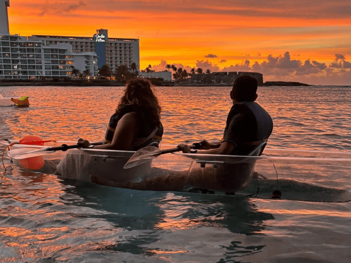 Two people in a transparent kayak on a sunset-lit ocean, near a hotel.