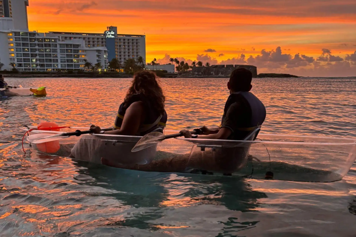 Two people in a transparent kayak on a sunset-lit ocean, near a hotel.