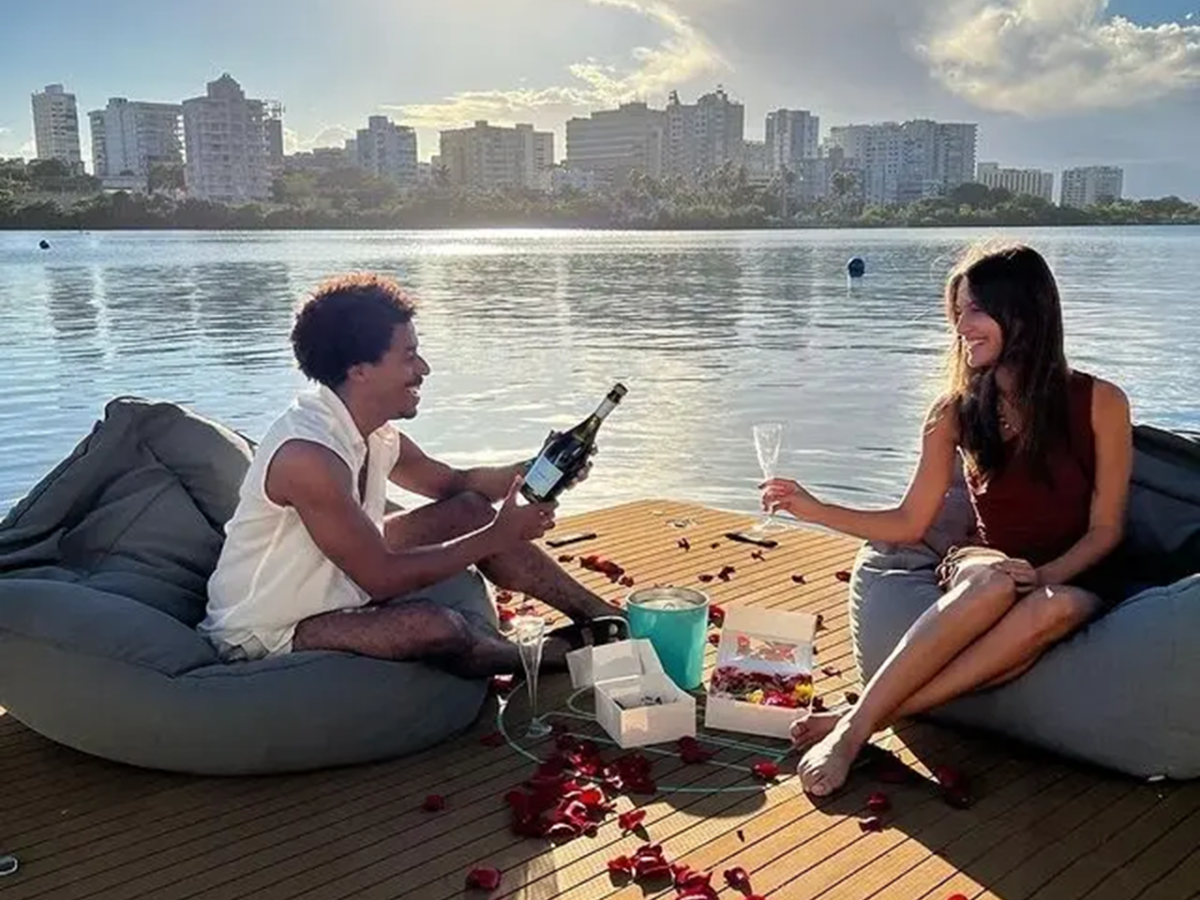 Couple on dock with wine and rose petals by a lake, city skyline in background.