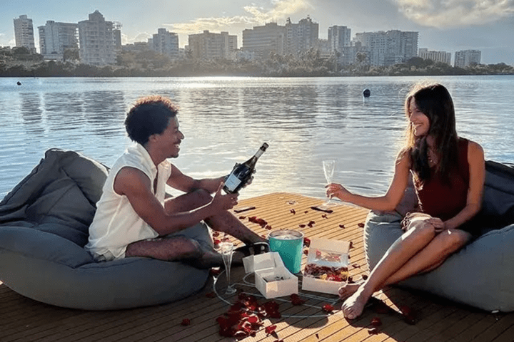 Couple on dock with wine and rose petals by a lake, city skyline in background.