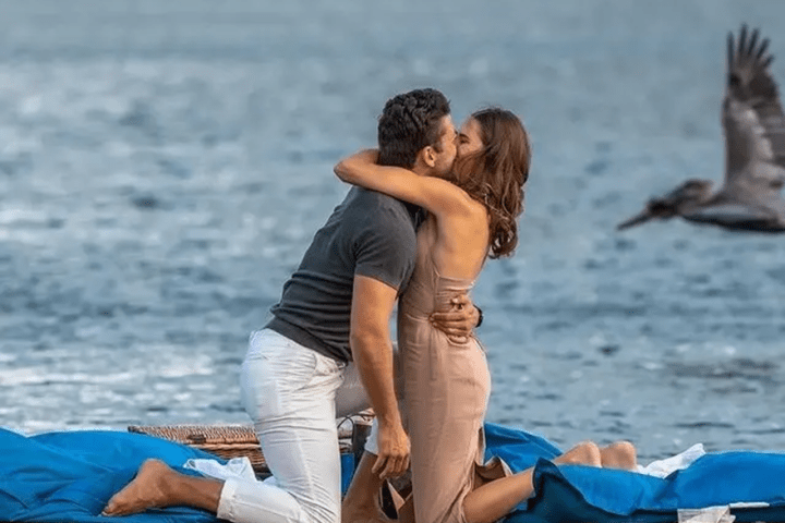 Couple kissing on raft, bird flying by, and green shoreline in background.