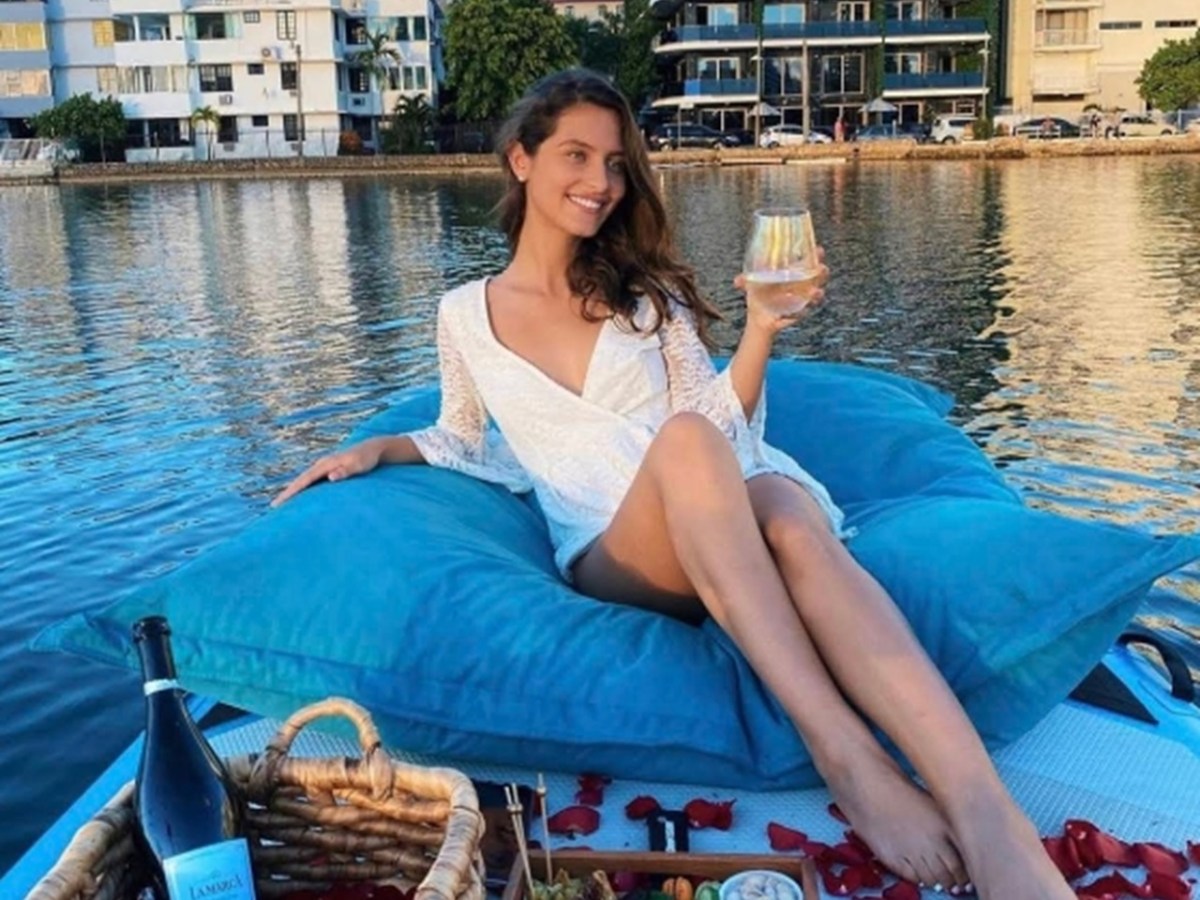 Woman relaxing on boat with wine, surrounded by picnic items on a waterway.