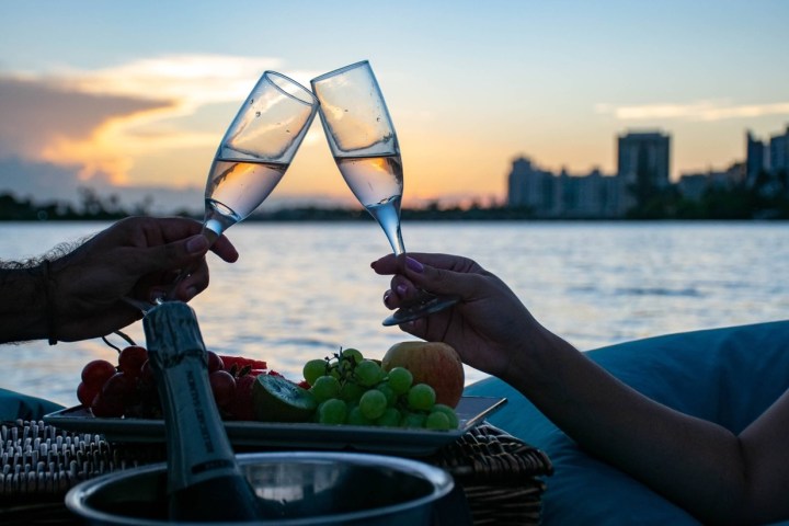 Two people clinking champagne glasses on a waterfront at sunset.