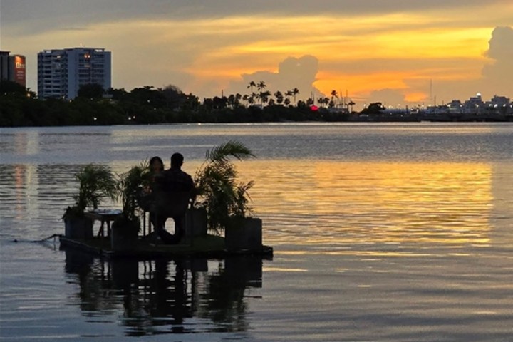 Two people on a raft with plants, silhouetted against a sunset over a calm lake.