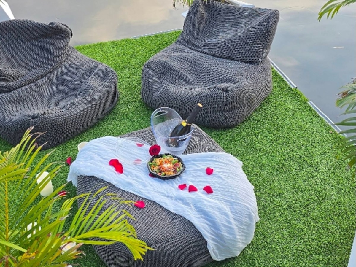Outdoor seating with gray cushions, a table with champagne, salad, and red petals on green grass by water.
