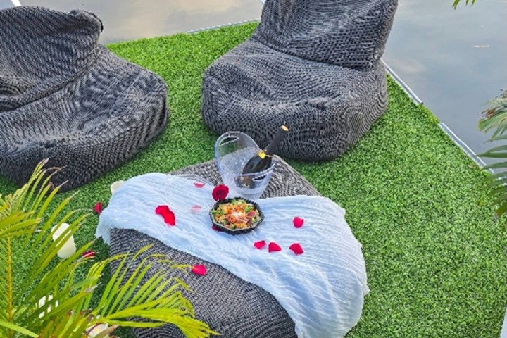 Outdoor seating with gray cushions, a table with champagne, salad, and red petals on green grass by water.