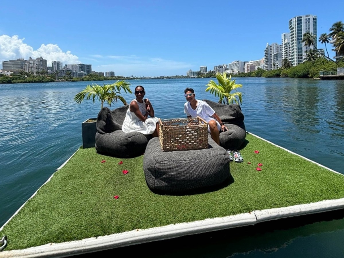 Two people sitting on floating platform with plants and cityscape view.
