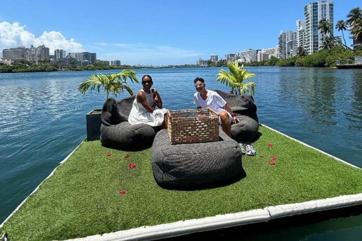 Two people sitting on floating platform with plants and cityscape view.