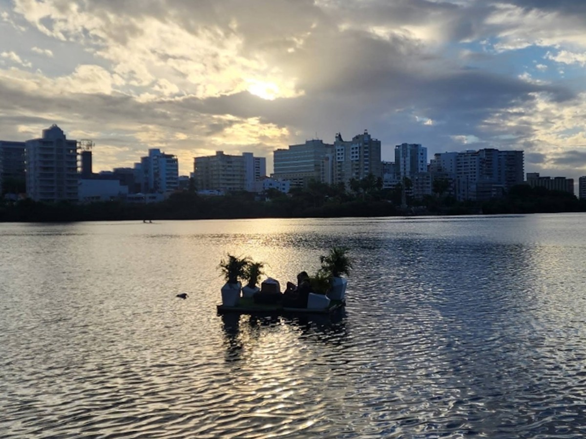 People on a small raft with plants on a lake at sunset, city skyline in background.