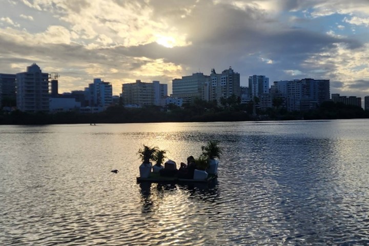People on a small raft with plants on a lake at sunset, city skyline in background.