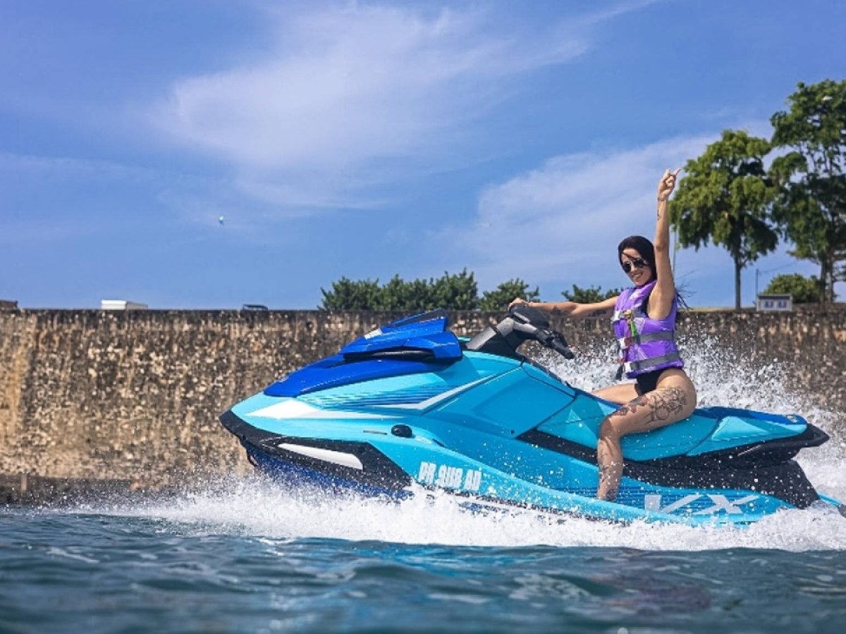 Person on a blue jet ski with raised arm, water splashing, stone wall and trees in background.