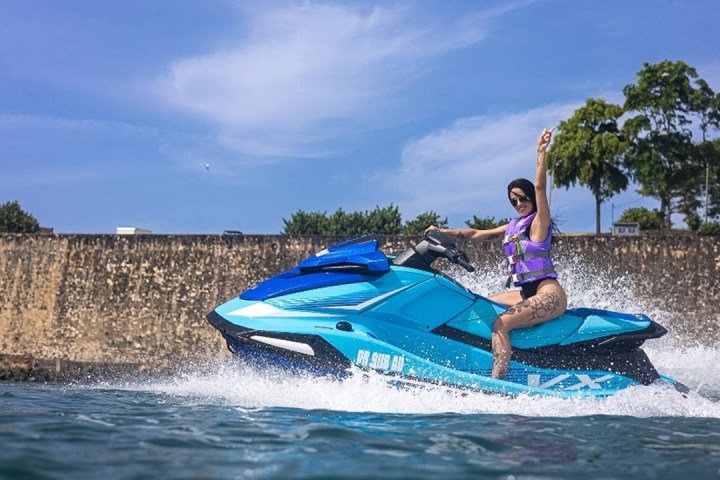 Person on a blue jet ski with raised arm, water splashing, stone wall and trees in background.
