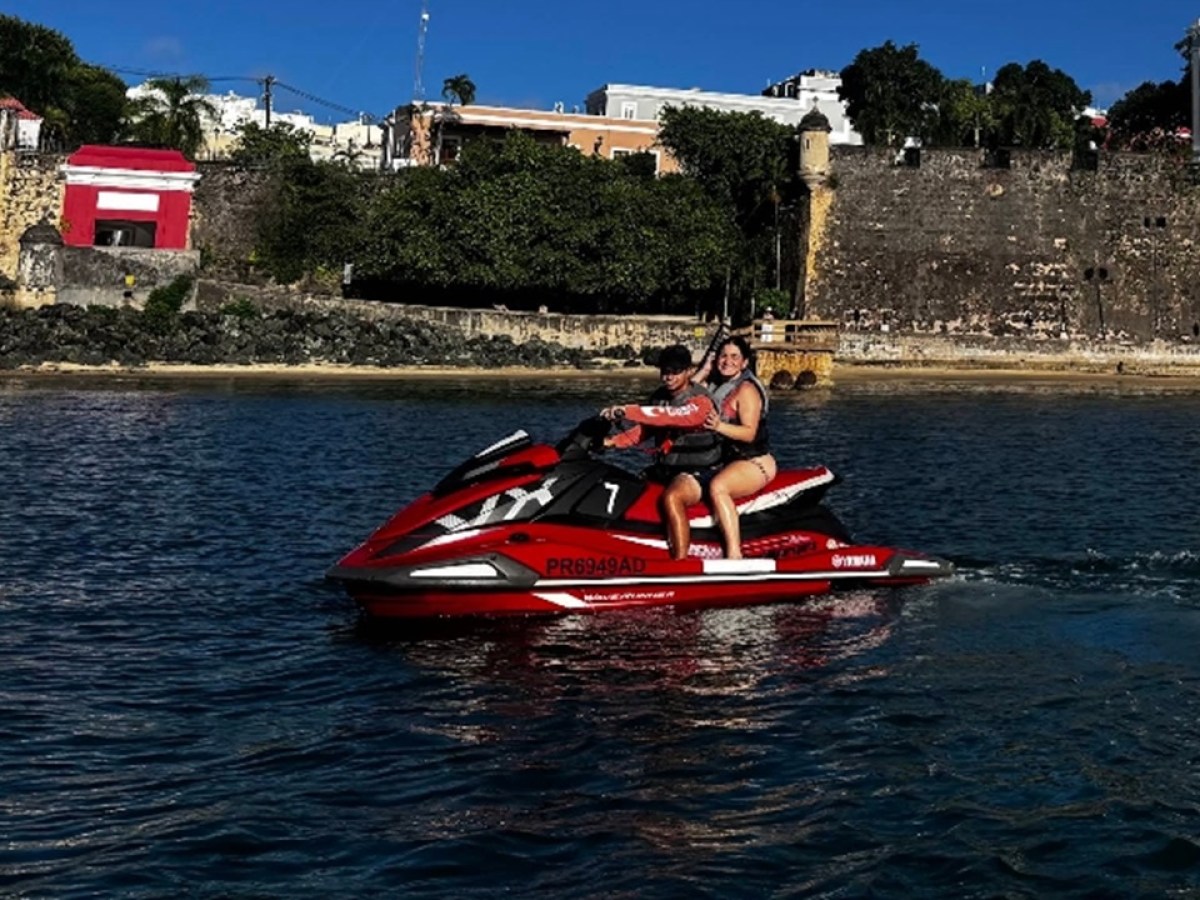 Two people on a red jet ski in front of an old stone building and trees.