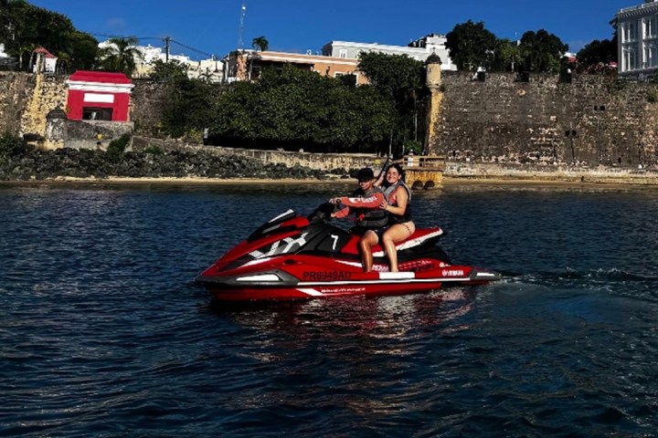 Two people on a red jet ski in front of an old stone building and trees.