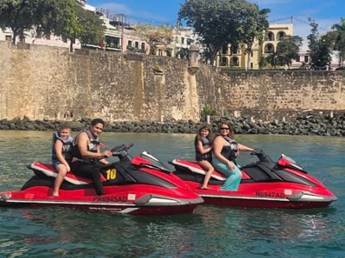 Two people riding red jet skis with a child each, near a historic stone wall.