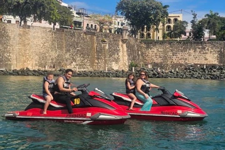 Two people riding red jet skis with a child each, near a historic stone wall.