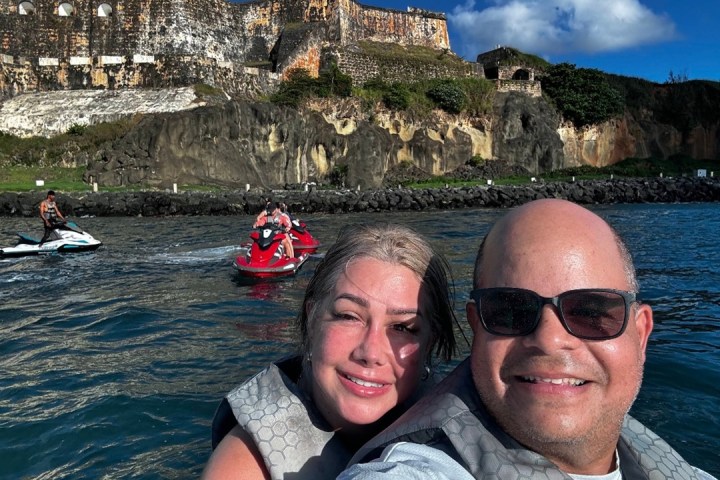 Two people smiling on jet skis with a historic fort in the background.